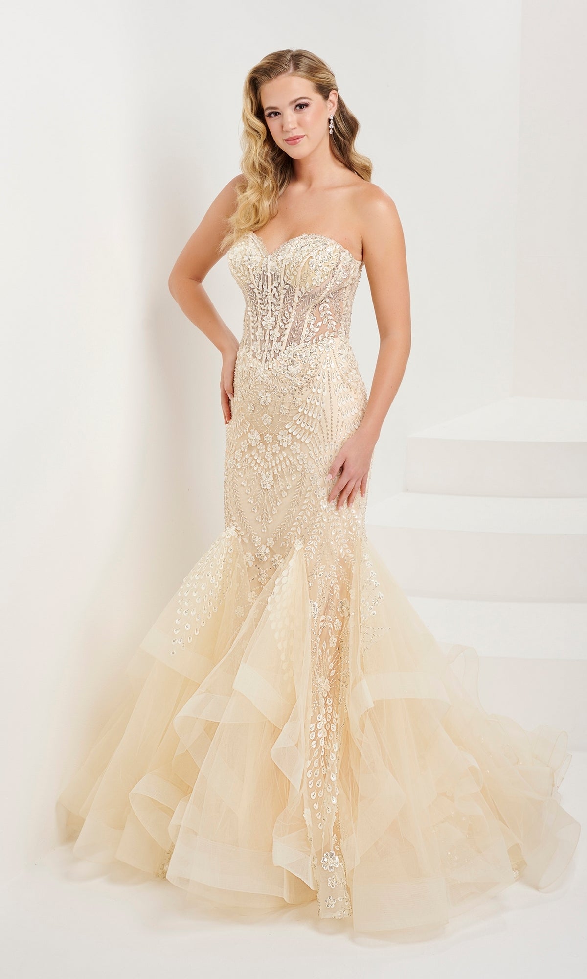 A woman stands against a white background in the Tiffany Strapless Beaded Mermaid Prom Dress 16103, a beige gown with beaded floral designs, sheer corset bodice, intricate beadwork, and a layered tulle skirt.